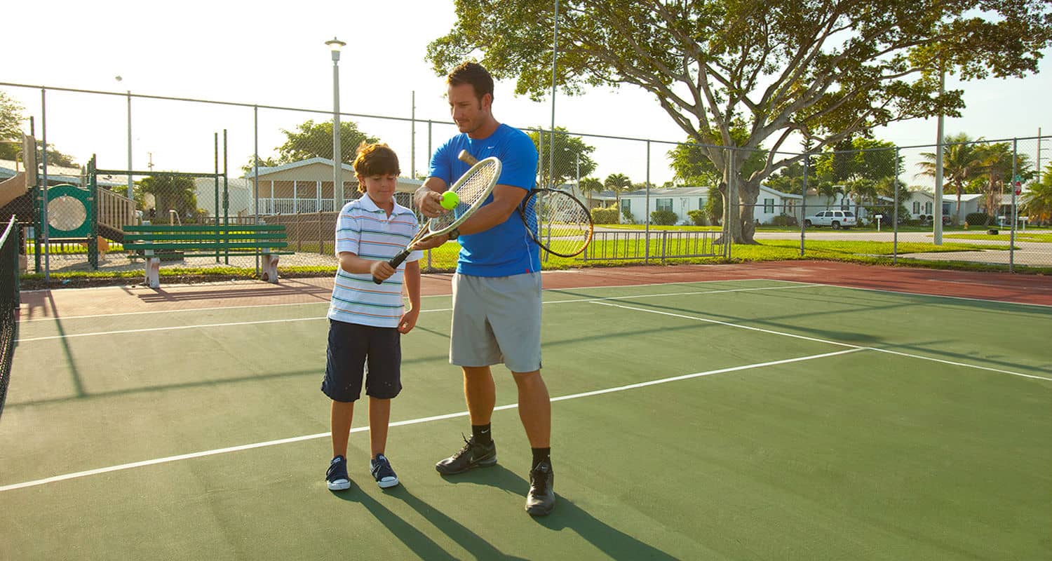 Rexmere Village tennis courts with a child’s playground in the background. A tennis teacher is giving a young man pointers on his grip.