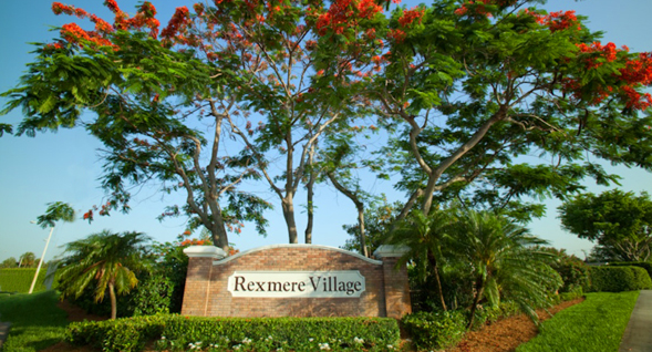 Brick and limestone entrance and sign to Rexmere Village, surrounded by flowers, trees and small palm trees and beautifully kept hedges.